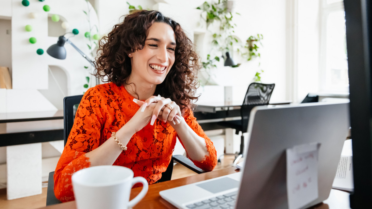A transgender woman smiling while video calling a business partner using her laptop at the office.